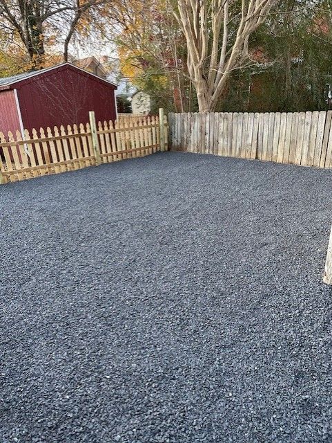 A backyard covered in dark grey gravel, surrounded by a wooden picket fence and a taller privacy fence, with a red shed.
