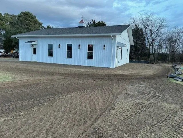 A white, single-story building with a dark roof and a cupola sits on a freshly graded dirt lot.