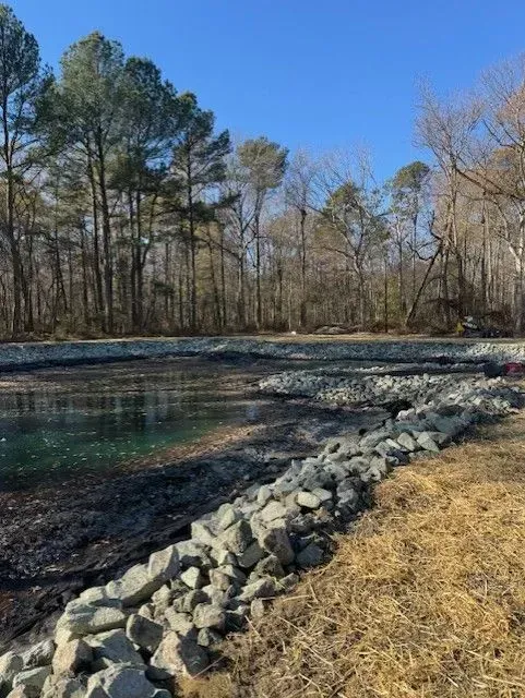 A man-made pond with rocky embankments, bordered by dry grass and a forest of tall trees under a clear blue sky.