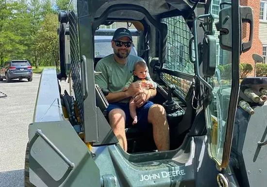 A smiling person wearing a cap and sunglasses holds a baby while sitting in the driver's seat of a John Deere skid steer.