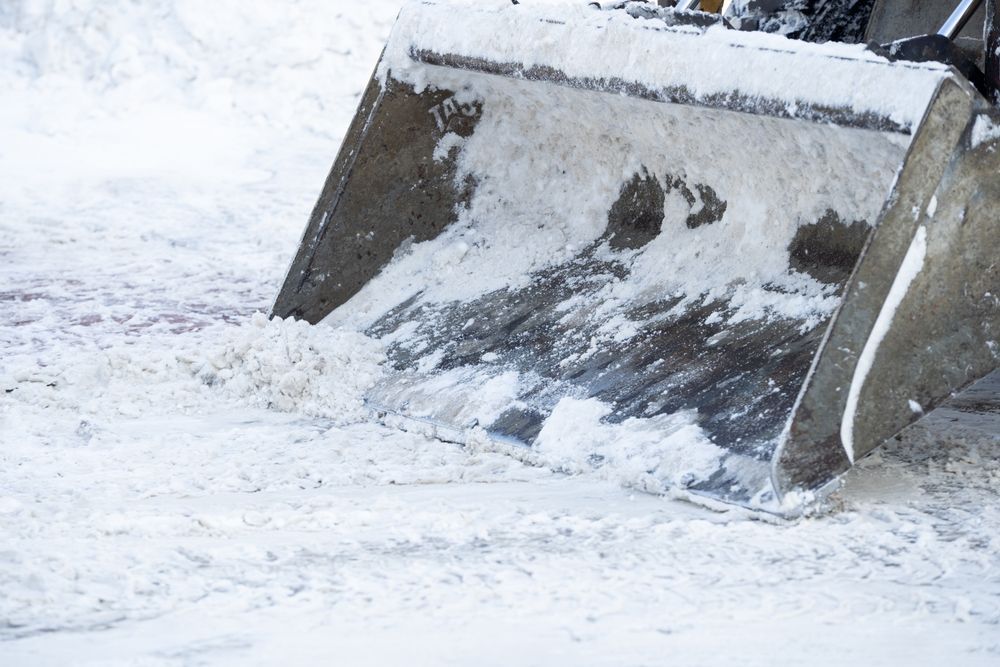 A close-up view of a metal snowplow blade clearing thick snow from a snowy surface.