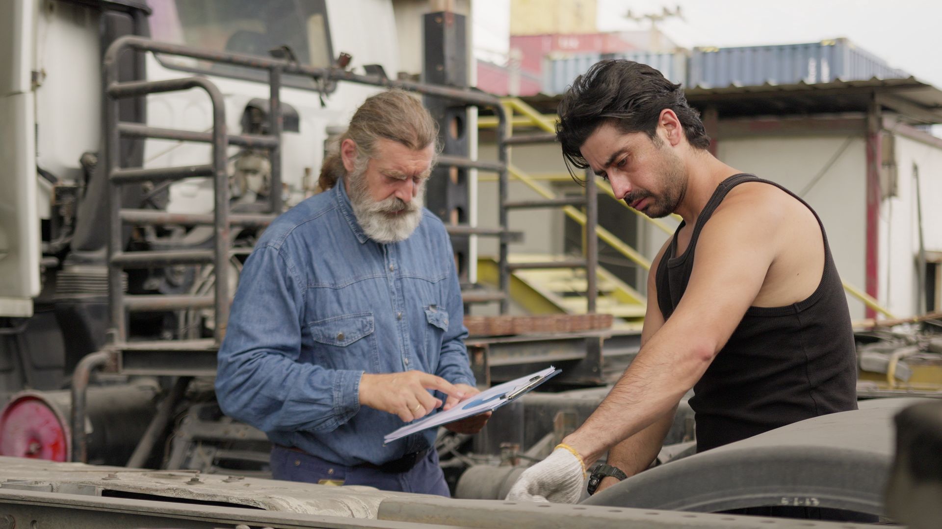 A senior mechanic supervises a younger technician performing truck maintenance.