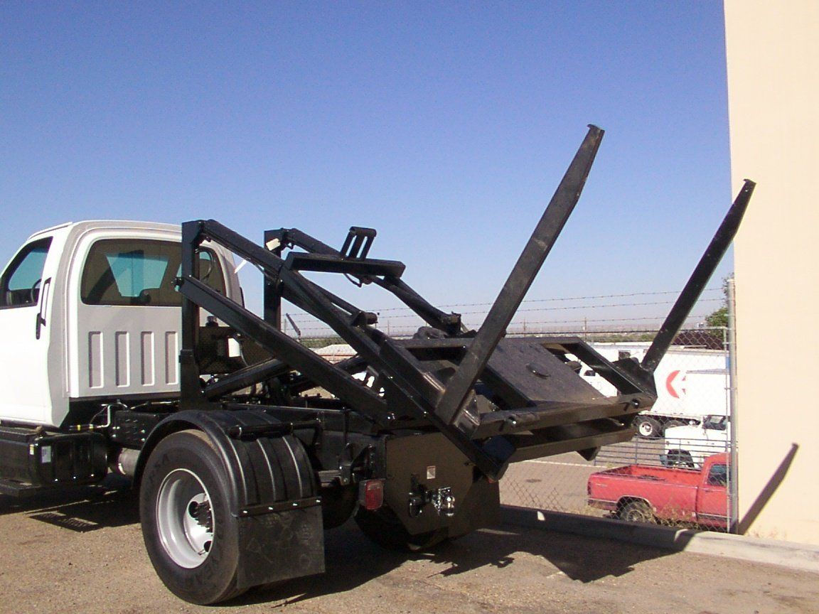 White truck chassis with black hook lift for hauling containers, parked outdoors in sunlight.