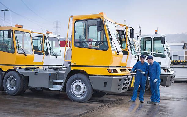 Two mechanics in blue coveralls reviewing a tablet next to a row of industrial, yellow and white tow trucks.