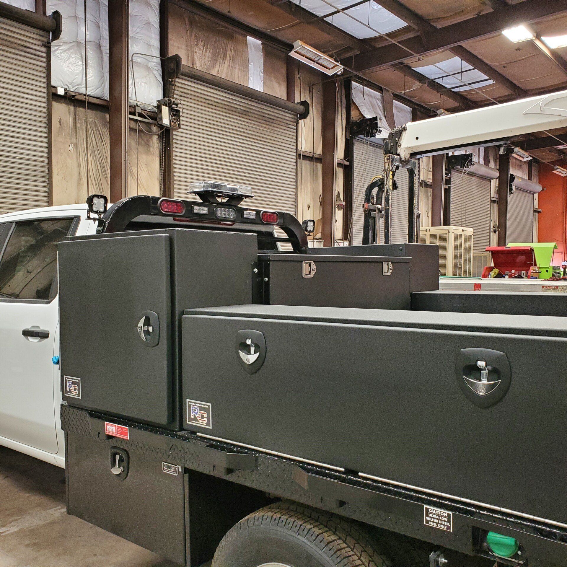 Black utility bed on a white truck in a warehouse. Includes toolboxes and a ladder rack.