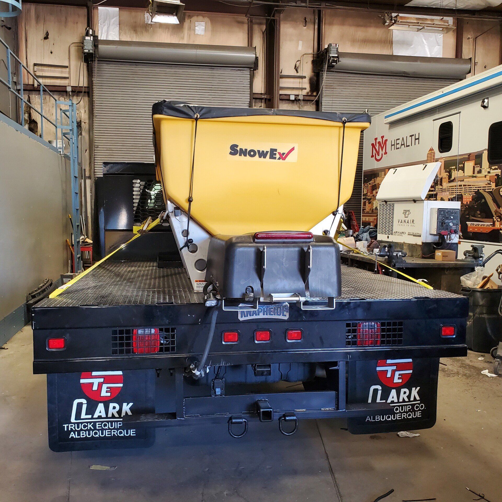 Yellow SnowEx salt spreader on a flatbed truck; Clark Truck Body logo visible.