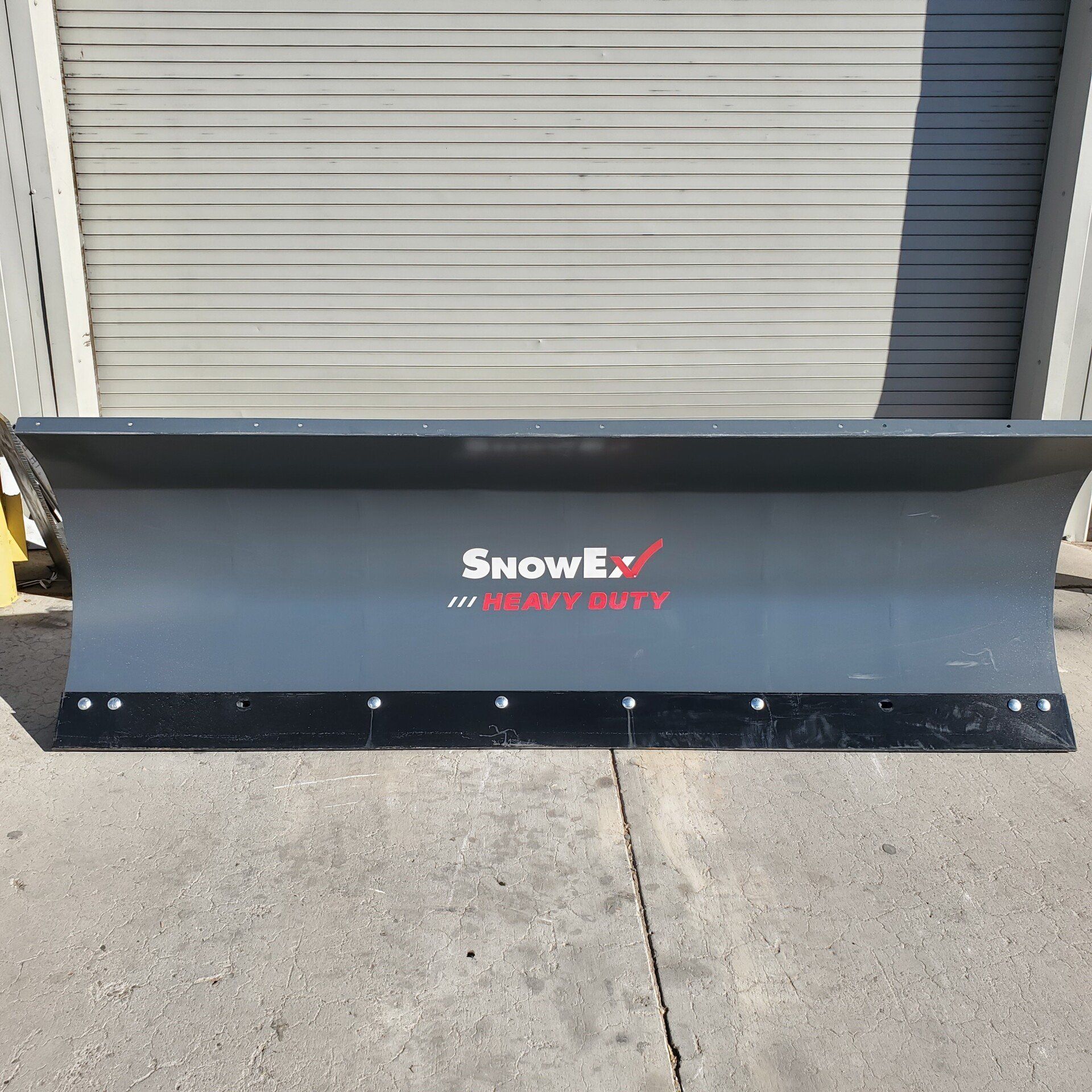SnowEx heavy-duty snow plow on a concrete surface. Gray blade with black cutting edge; red and white logo.