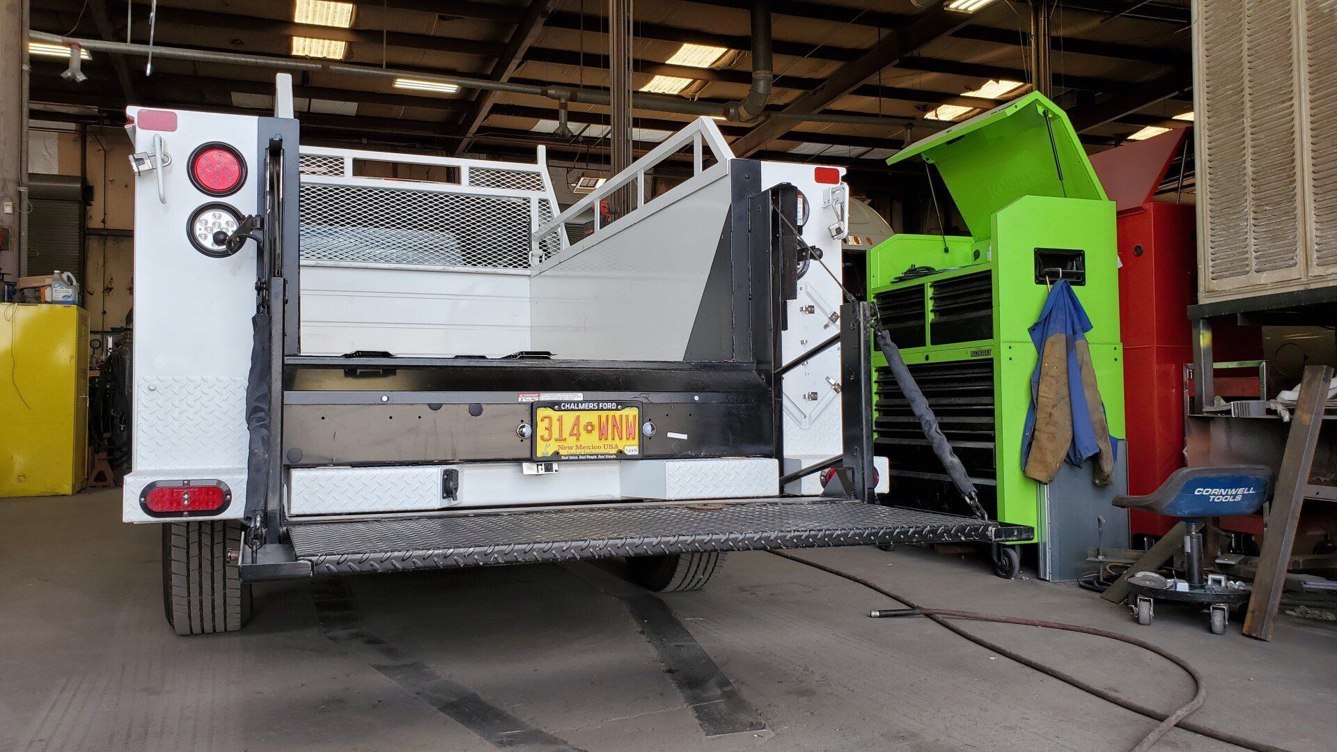 A utility truck with its tailgate down inside a workshop. Green toolbox to the right.
