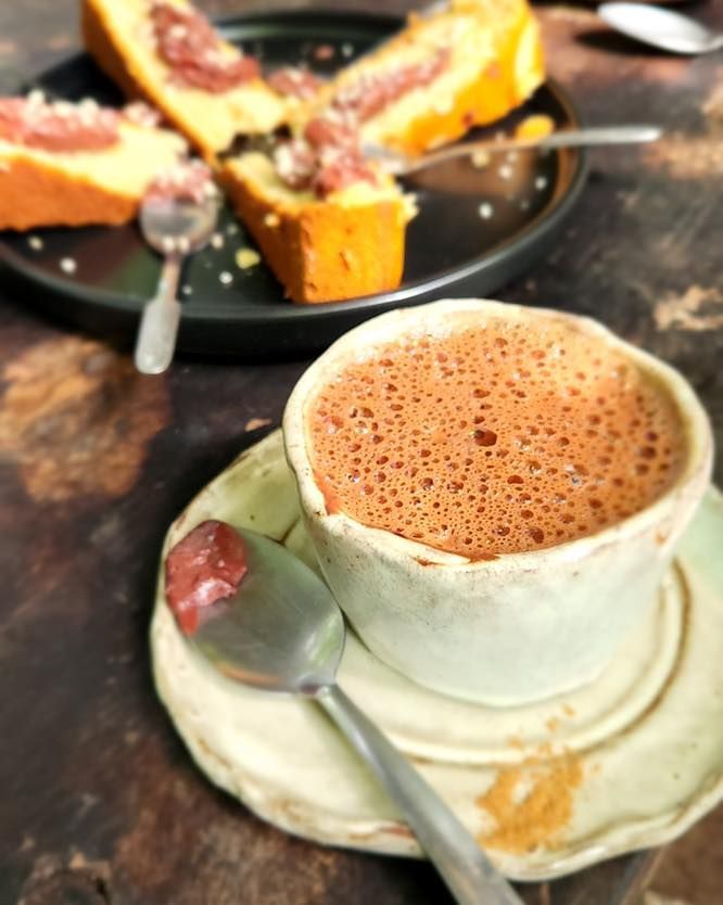 Foamy coffee in a white cup on a saucer, with cake slices blurred in the background.