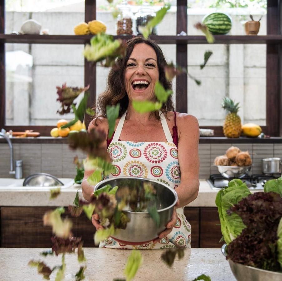 Woman in a kitchen tossing leafy greens into the air while smiling, with fresh produce on shelves behind her