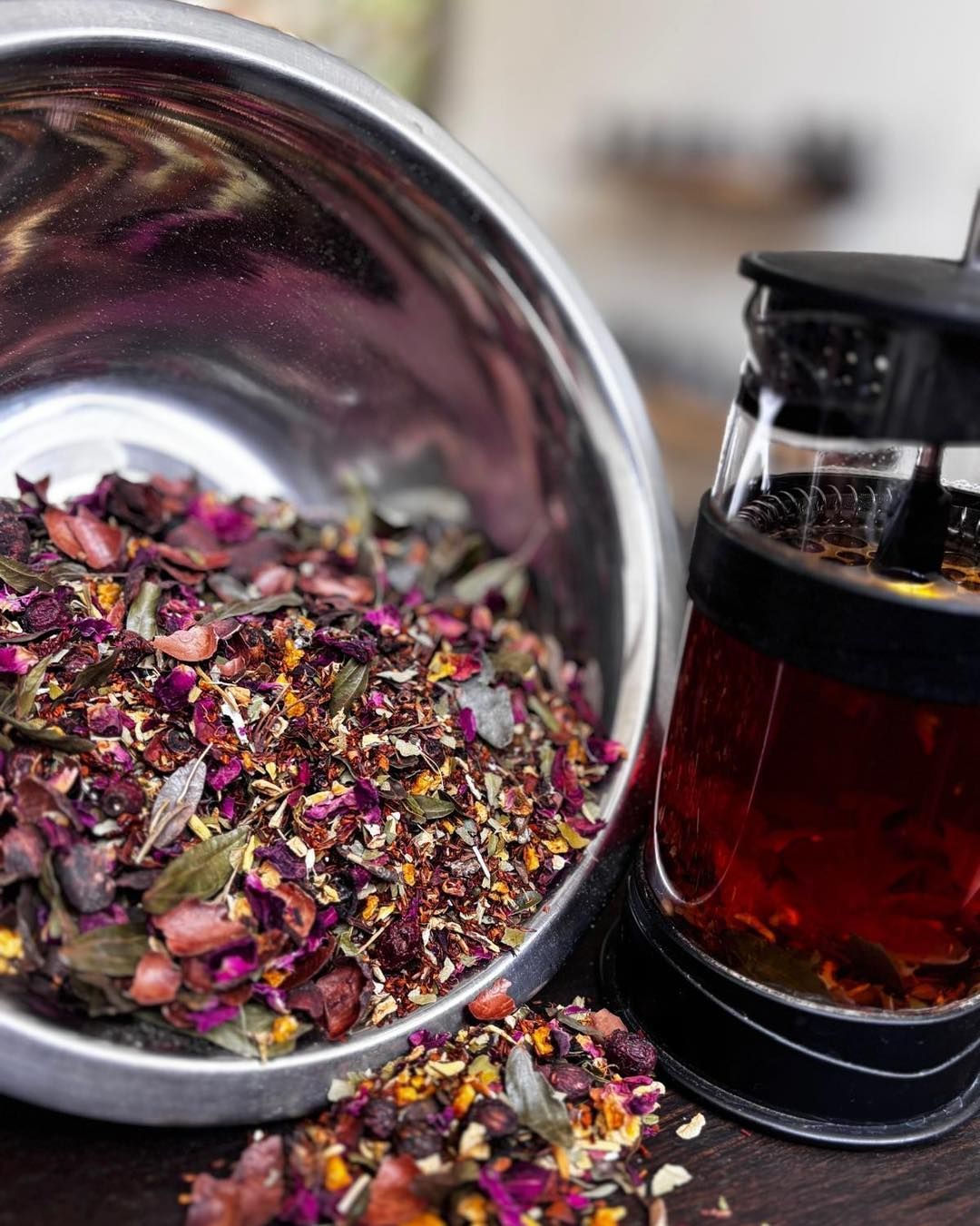 Loose dried flower petals in a metal bowl beside a small glass jar of red tea