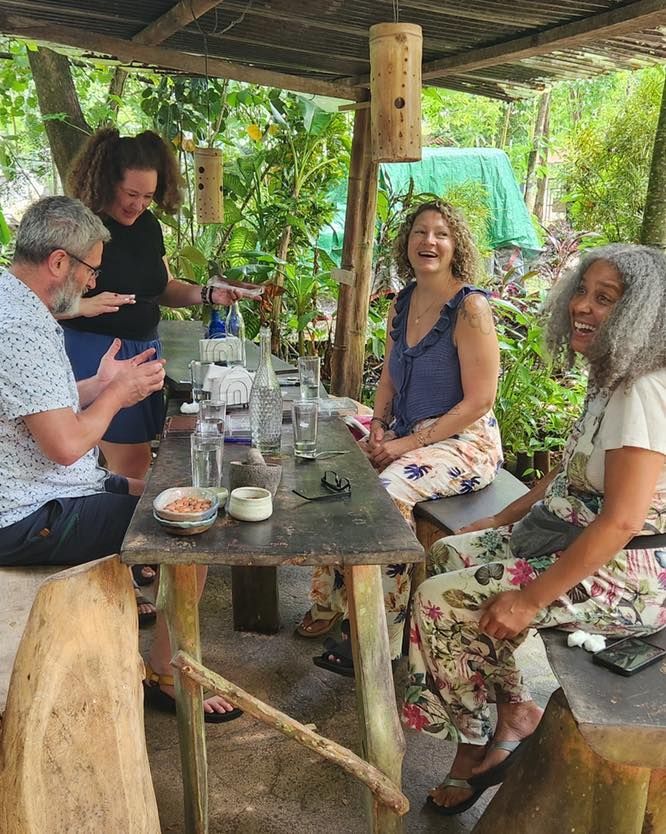 Four people chatting at a rustic outdoor table in a lush tropical setting.
