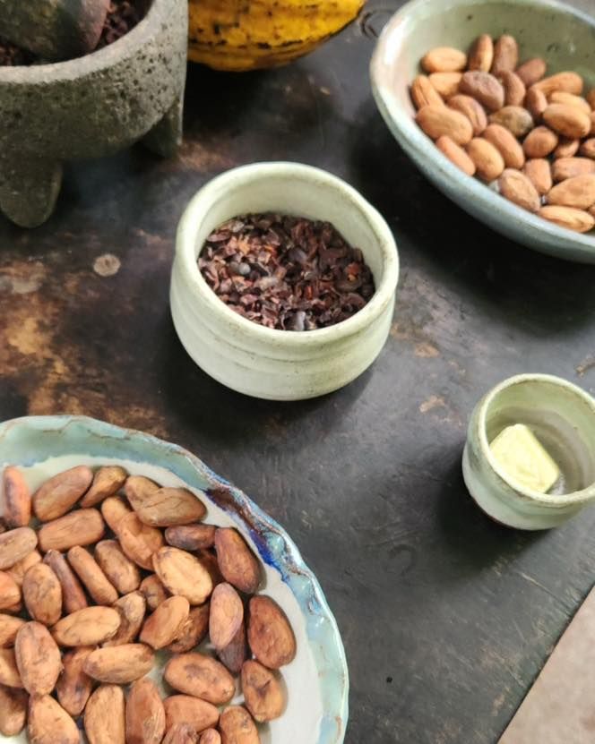 Bowls of cocoa beans and pods on a dark tabletop, with a small cup of cocoa nibs.