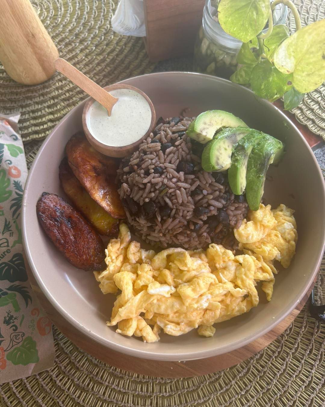 Breakfast bowl with scrambled eggs, rice, avocado, plantain, sausage, and yogurt on a woven table.