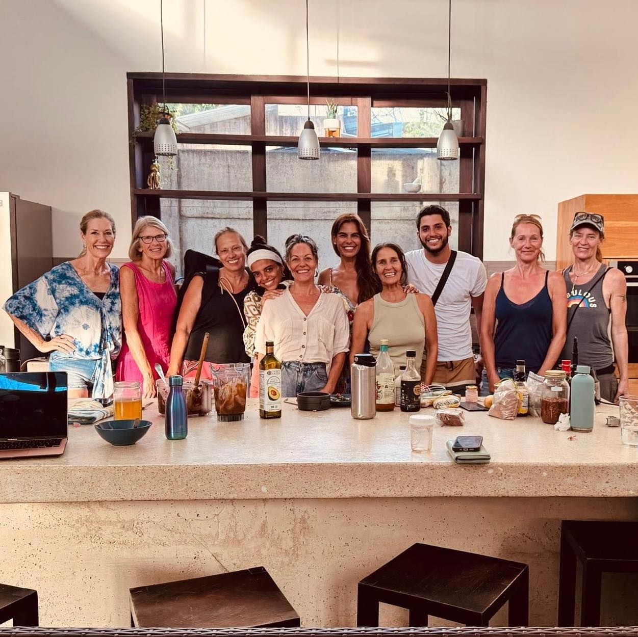 Group of people smiling together in a kitchen behind a counter, with windows and drinks visible.
