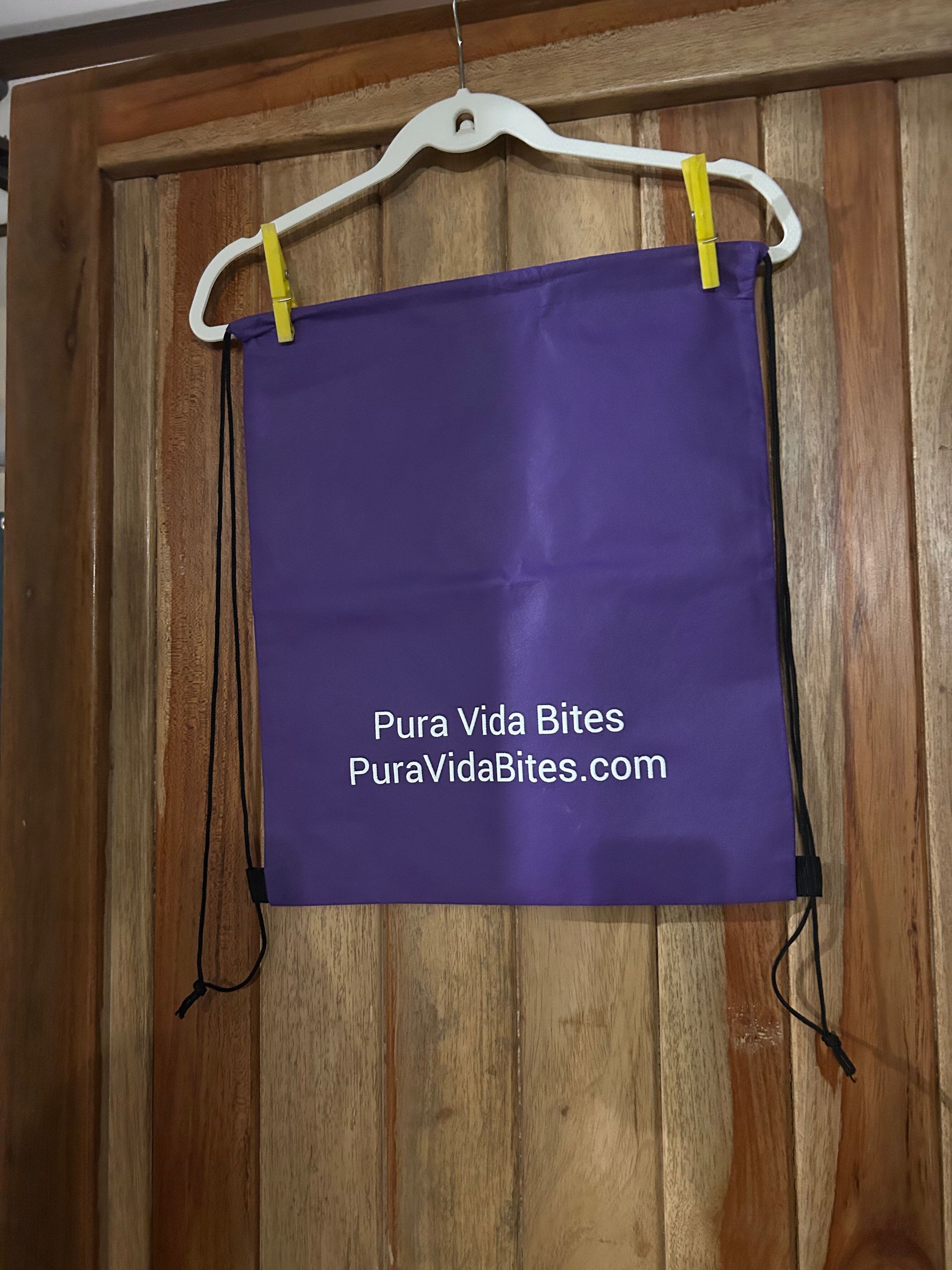 Purple drawstring bag hanging on a white hanger against a wooden door, labeled PuraVidaBites.com