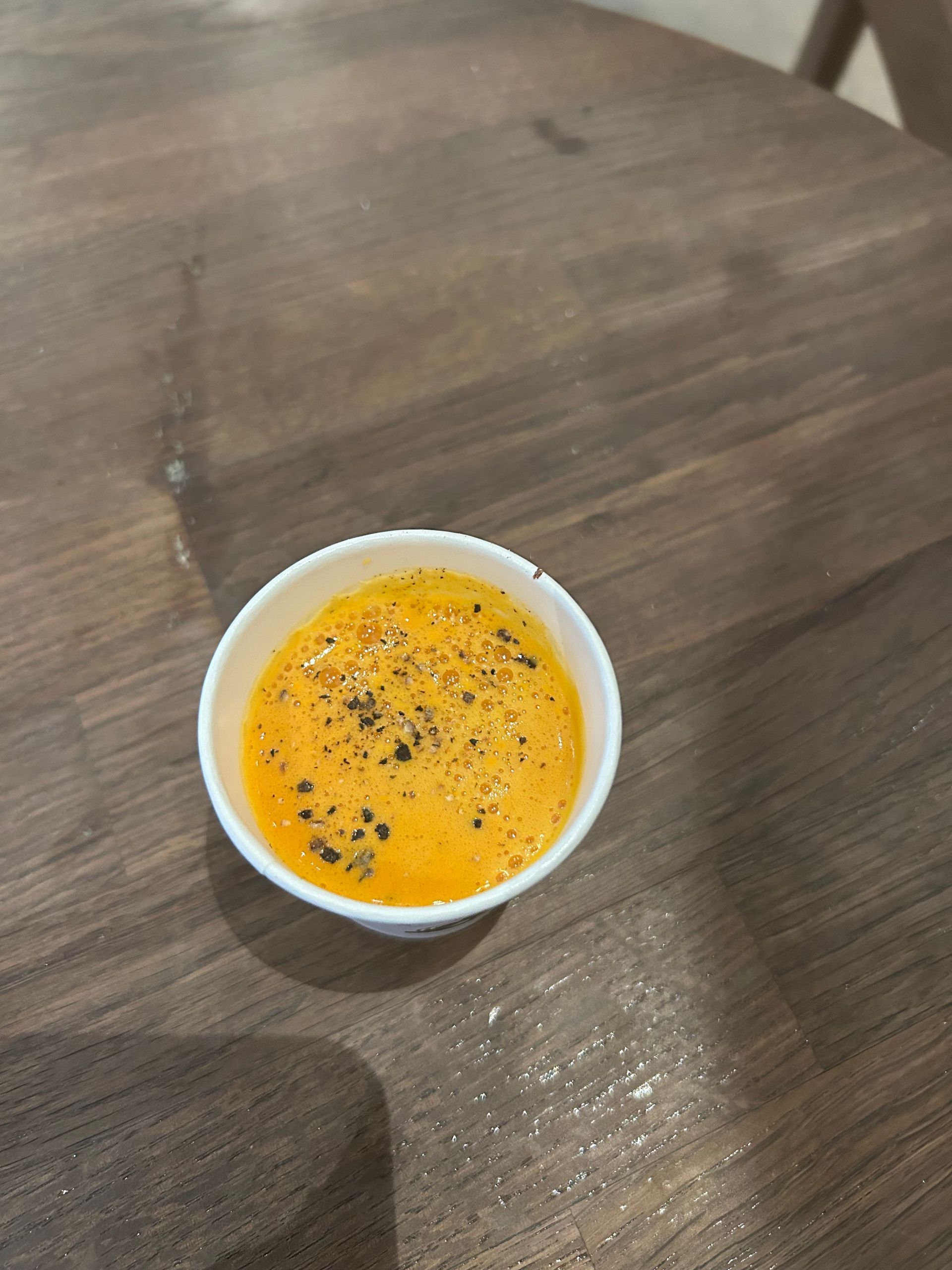 Small white cup of orange soup topped with black pepper on a wooden table.