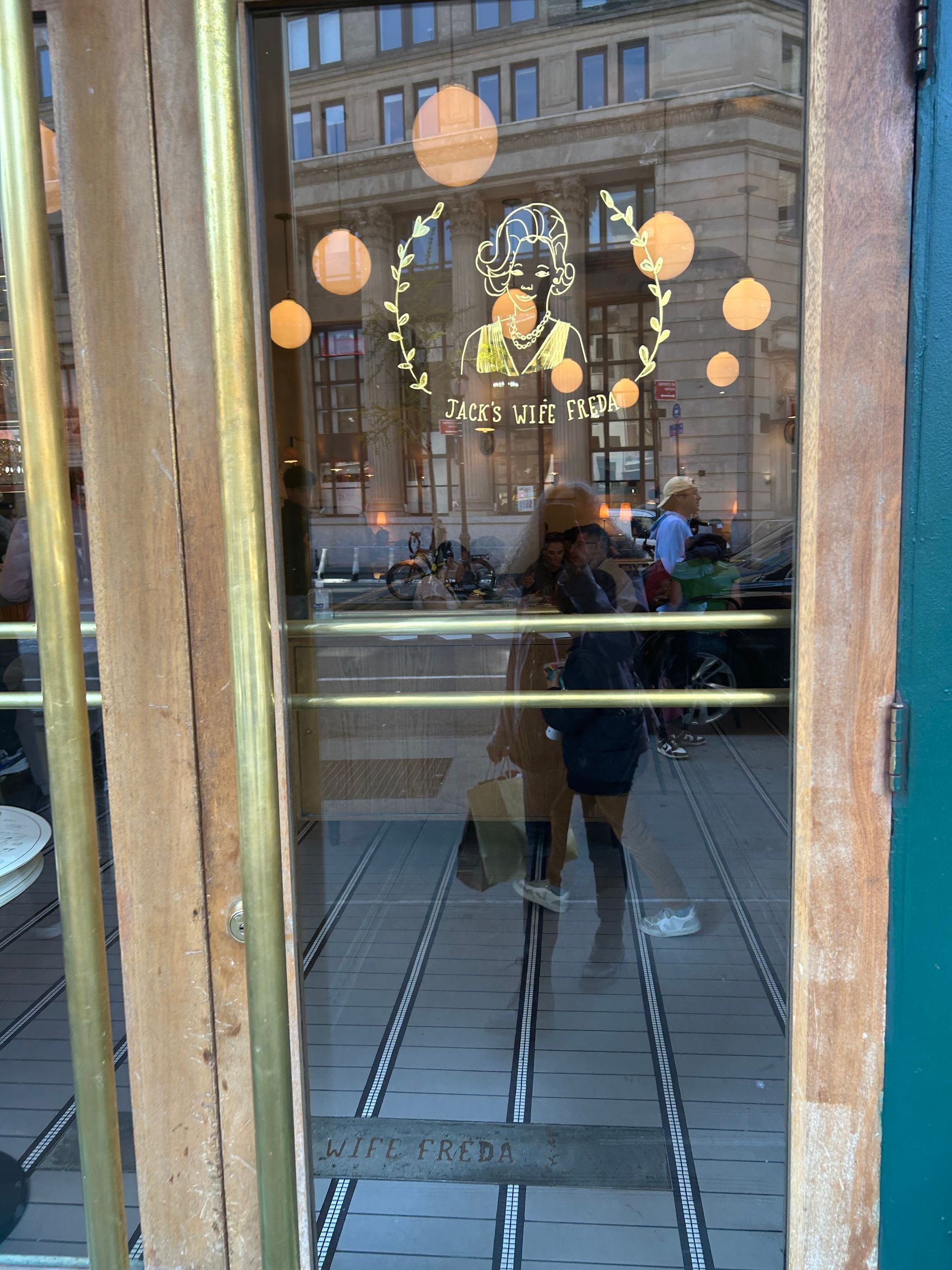 Person reflected in a glass storefront, with hanging lanterns and a brick building across the street.