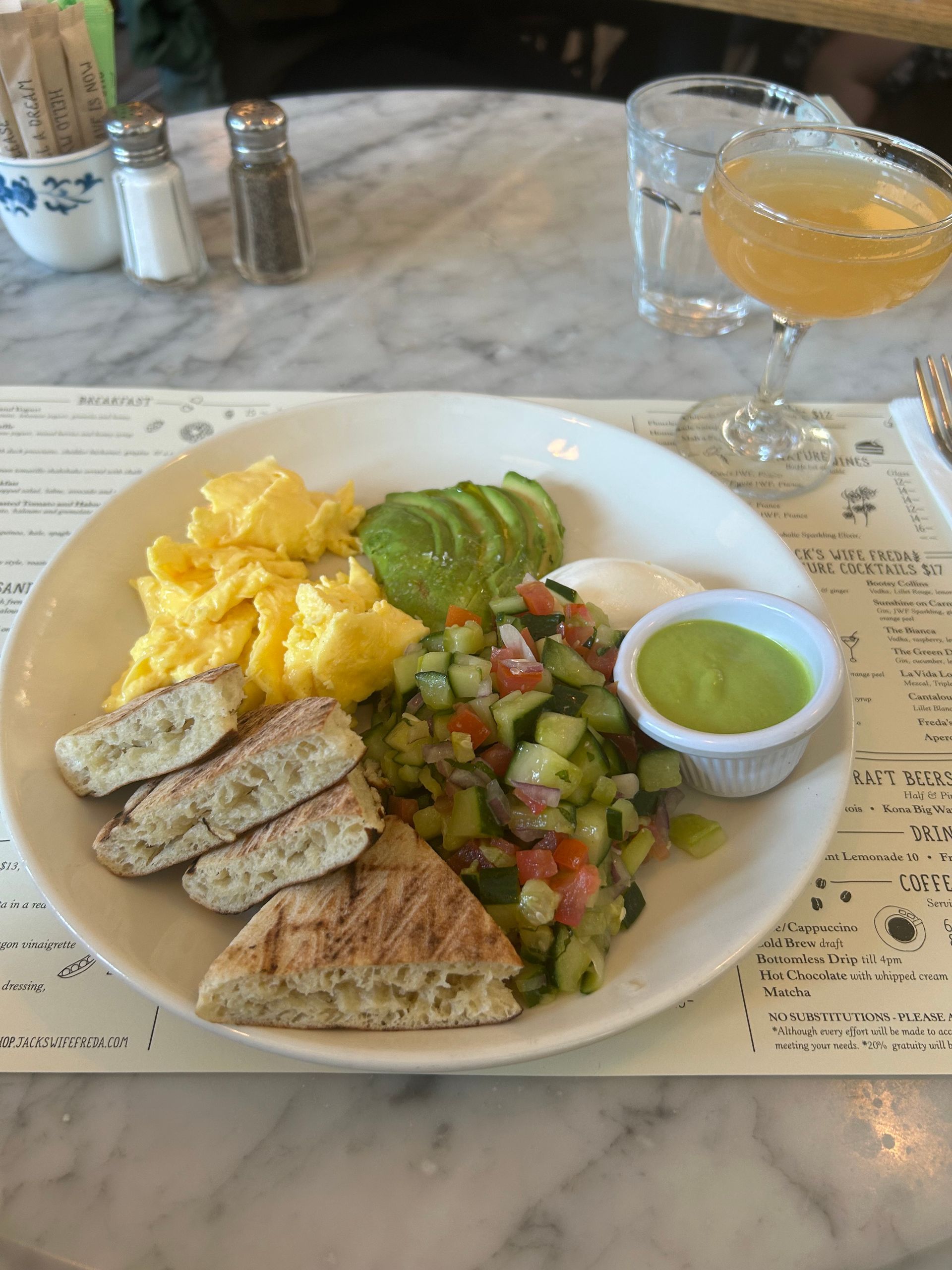 Brunch plate with scrambled eggs, avocado, salad and bread, beside a cocktail on a marble table.