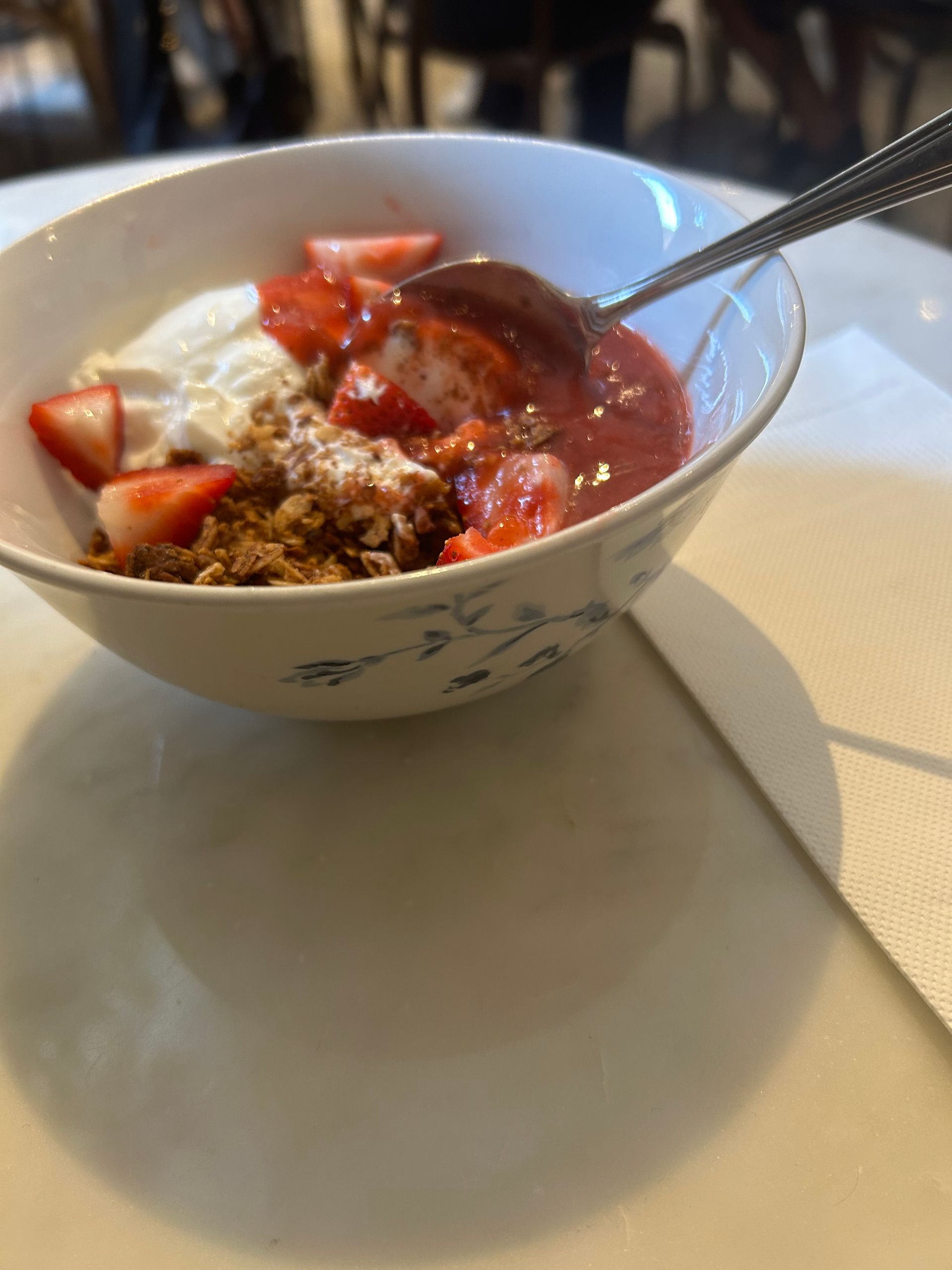 Bowl of yogurt topped with strawberries and granola, with a spoon on a table.