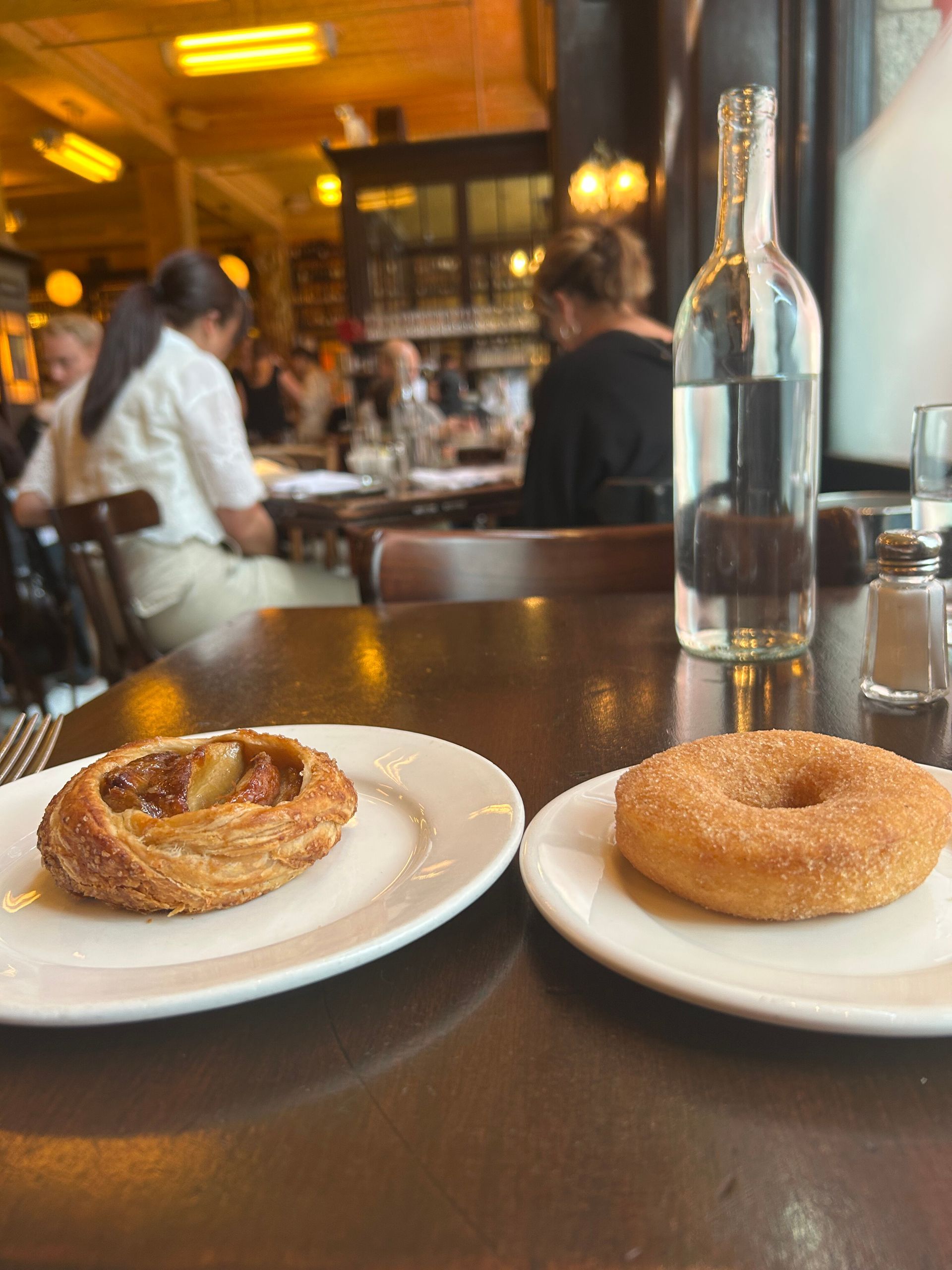Two pastries on white plates at a café table with a glass bottle in the center, blurred diners in the background