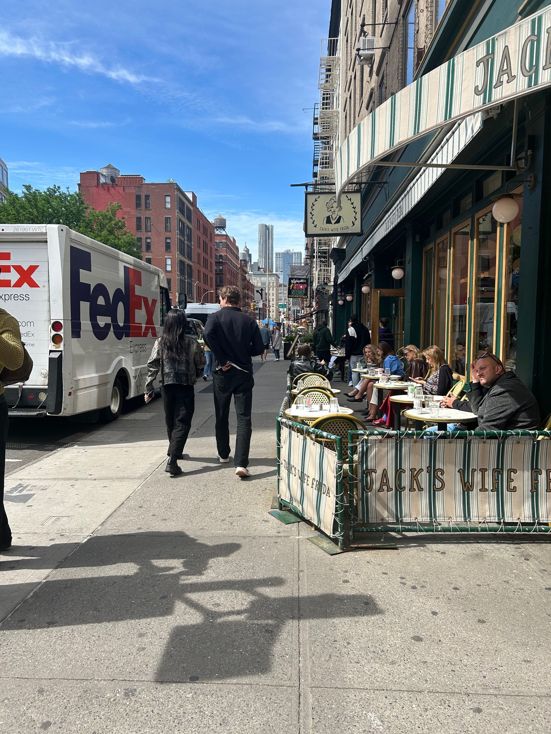 Sidewalk cafe on a sunny city street with pedestrians, parked trucks, and brick buildings.