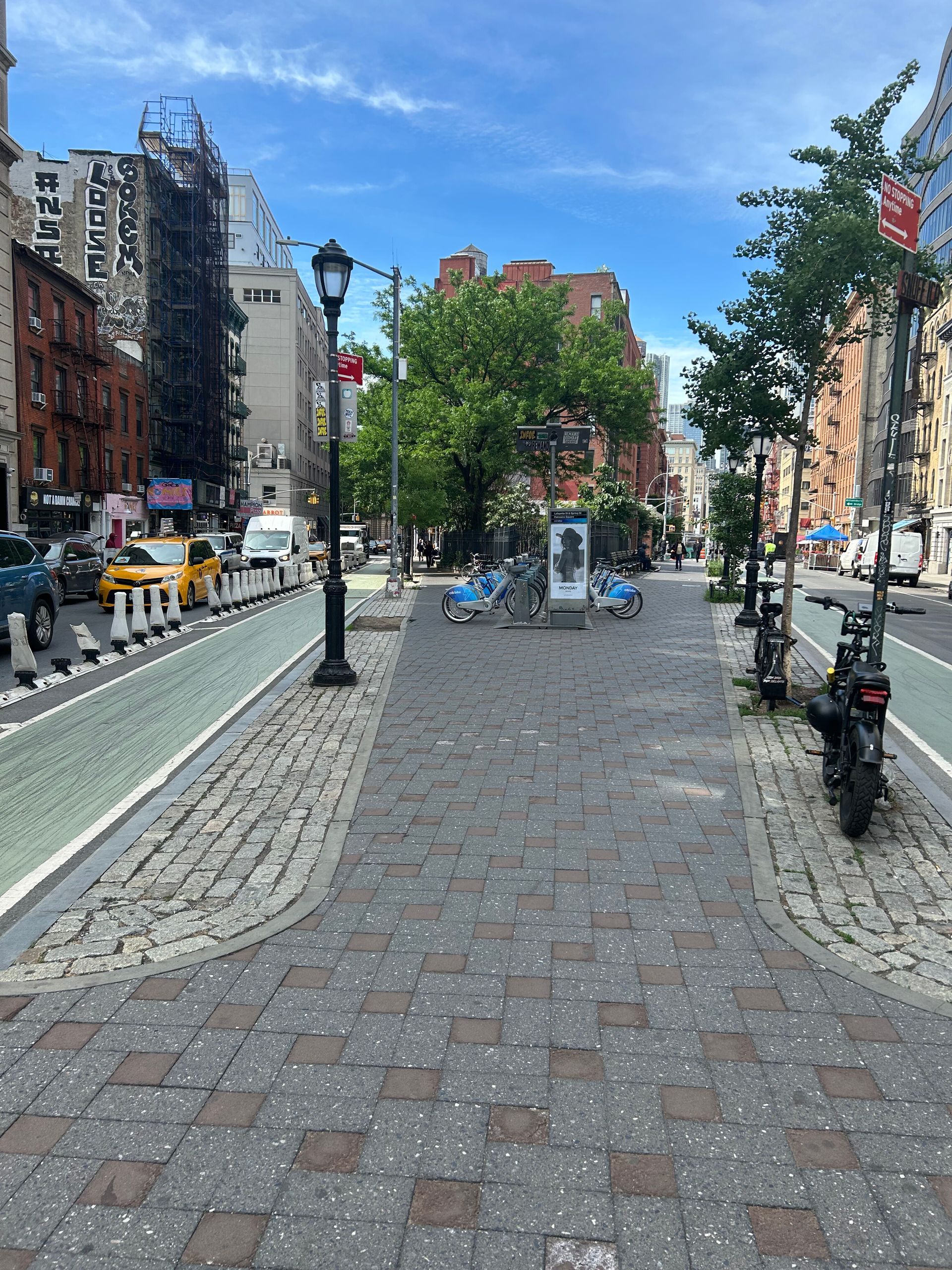 Urban street with cobblestone walkway, lampposts, trees, parked bikes, and buildings under a blue sky
