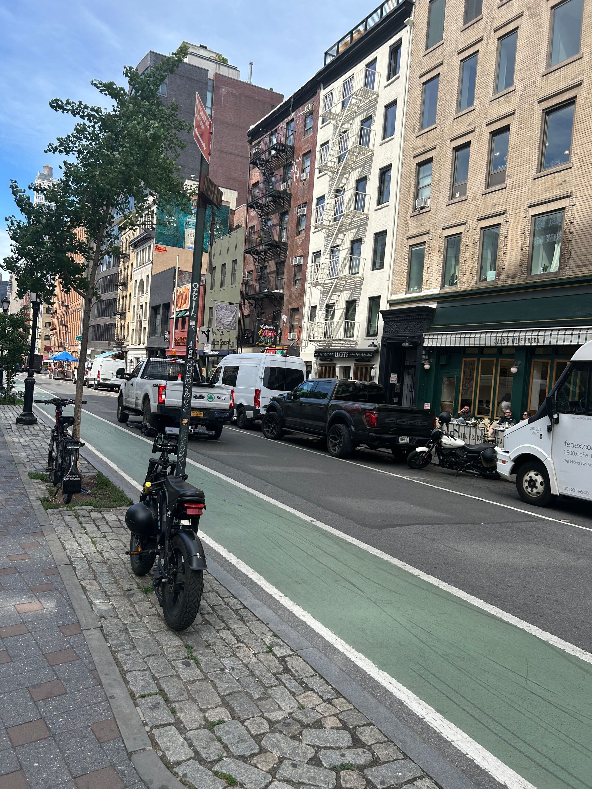 City street with parked cars, a bike lane, and brick apartment buildings along a tree-lined sidewalk.