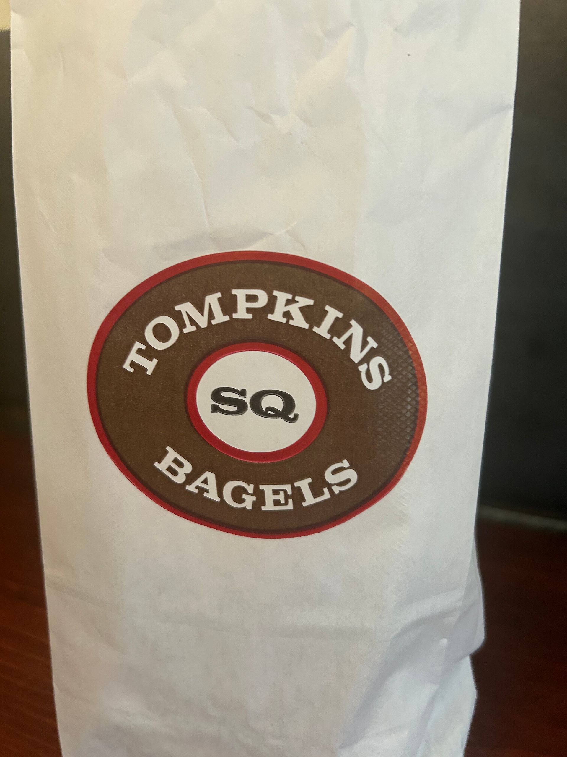White paper bag with a brown-and-red Tompkins Bagels logo and “SQ” in the center.