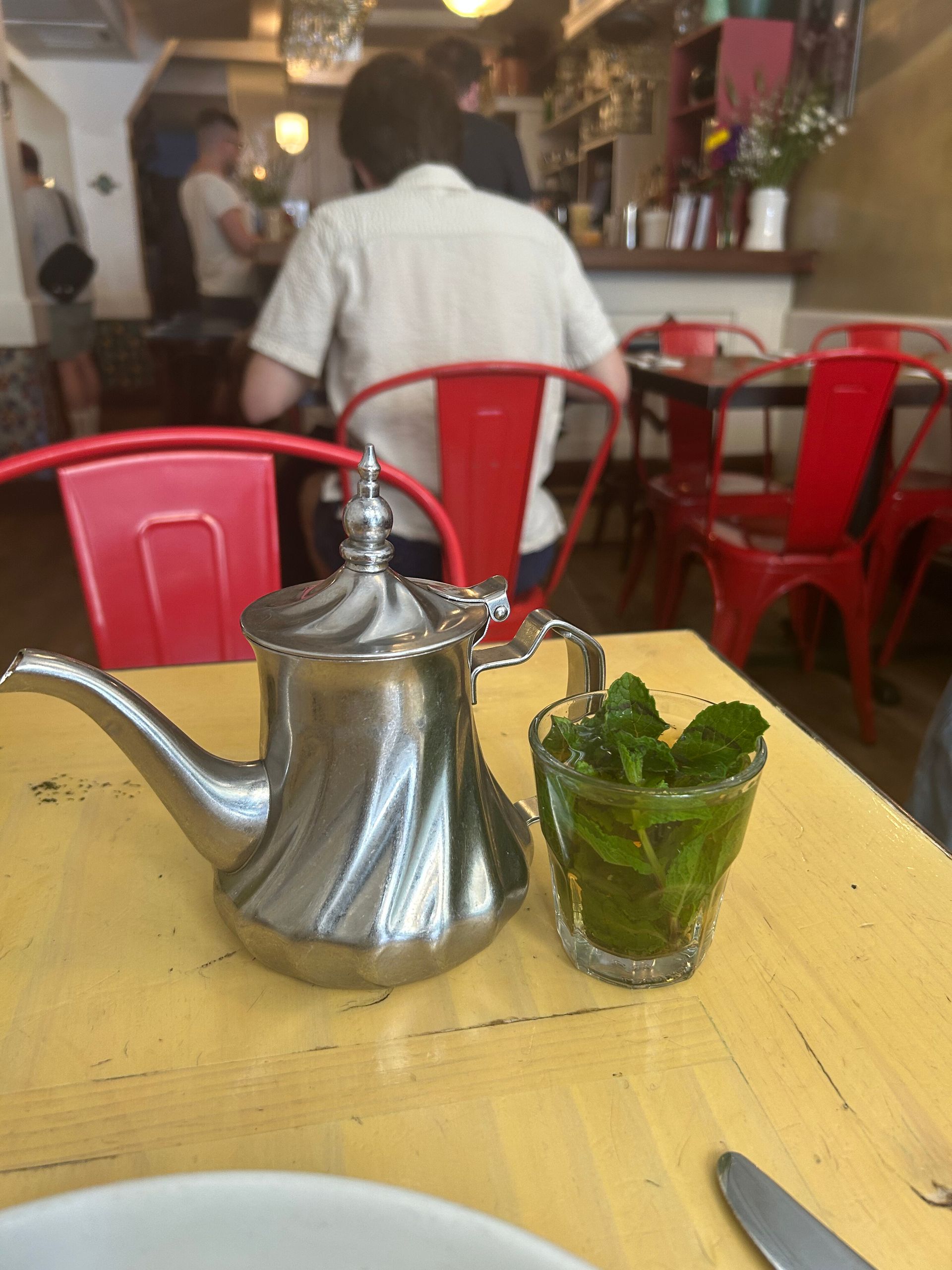 Tea set and mint drink on a yellow table in a café with red chairs and a blurred person in the background