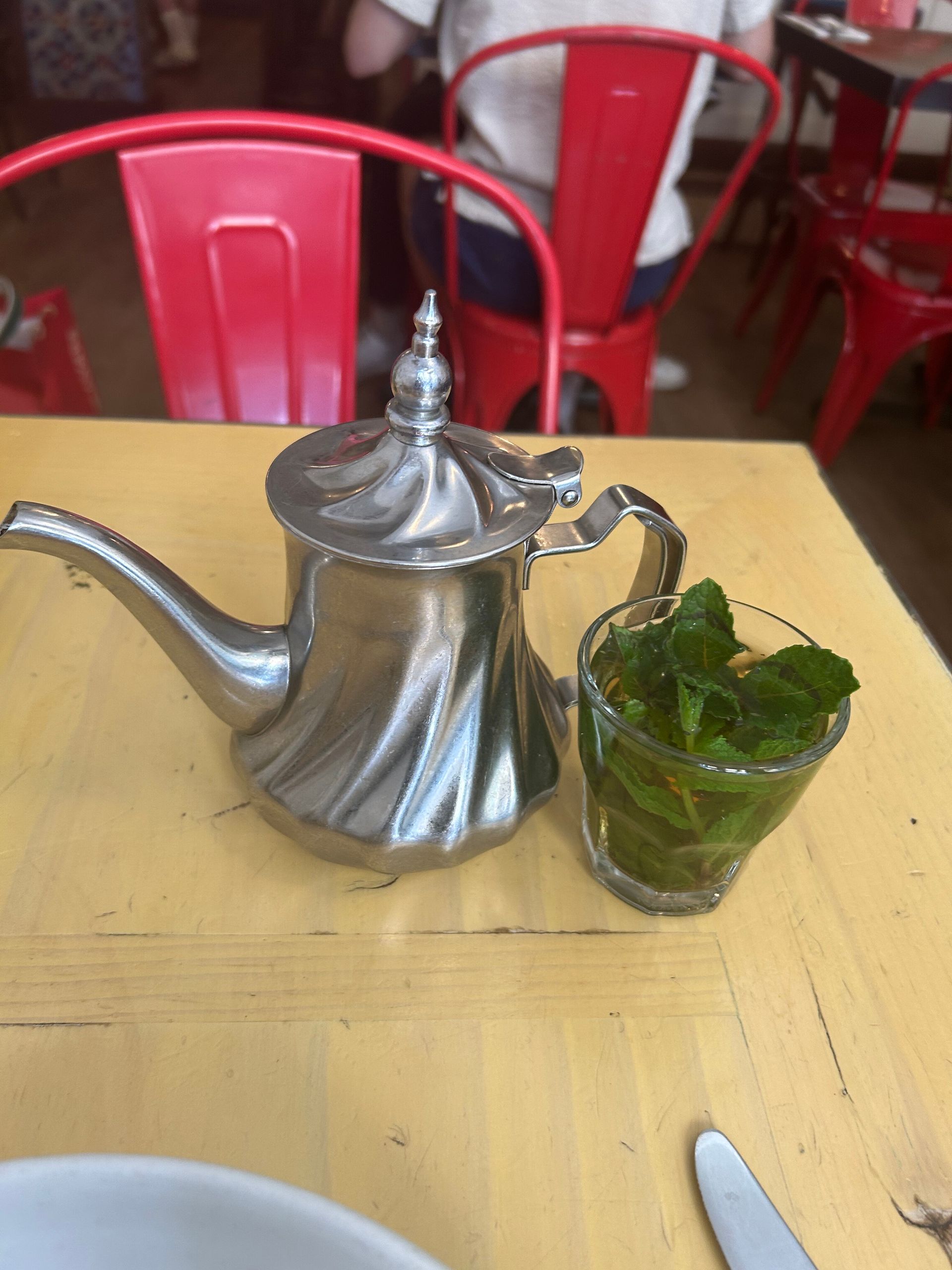 Silver teapot and glass of mint tea on a yellow table with red chairs in the background