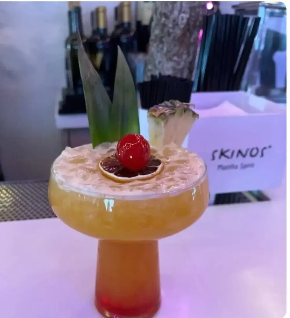 Tropical cocktail in a stemmed glass with cherry garnish on a table, with a Kinor menu in the background