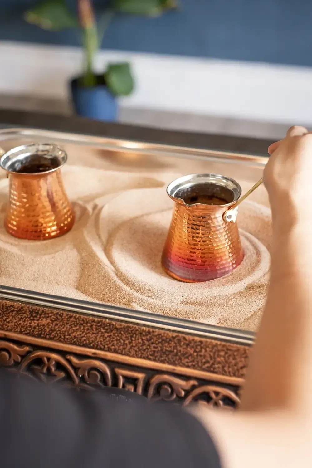 Two copper Turkish coffee cups on a decorative tray, with a hand reaching in from the right.