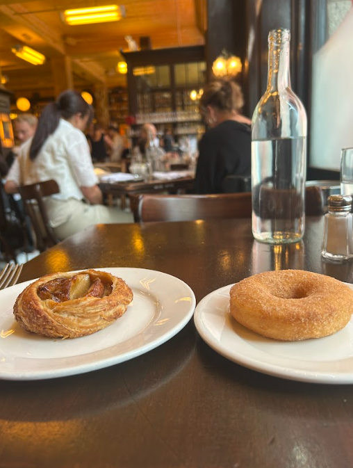 Two pastries on white plates at a café table, with a glass bottle and blurred diners in the background.