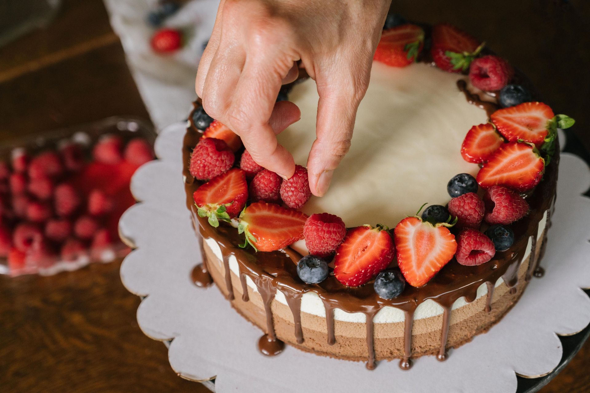 Hand placing strawberry slices on a chocolate-drizzled cake on a white cake board