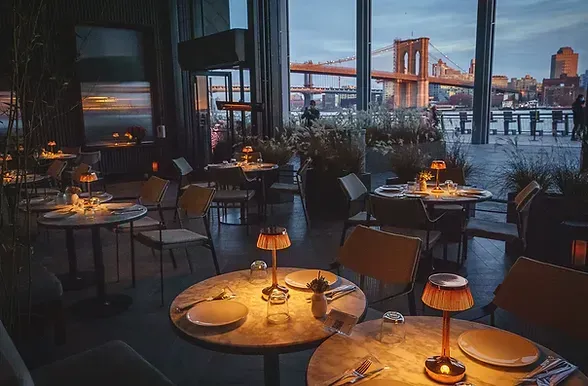 Dim riverside restaurant patio at dusk with lit tables and city skyline beyond.