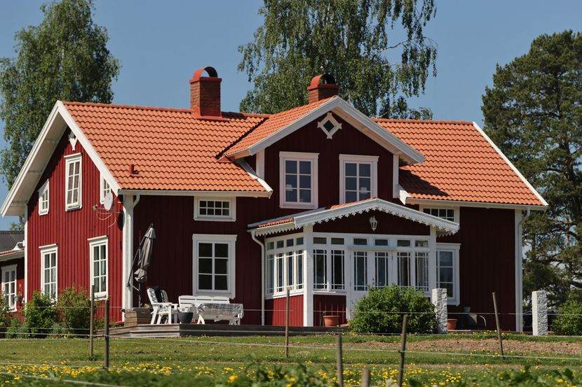 Red wooden house with white trim and orange roof in a grassy garden under clear sky