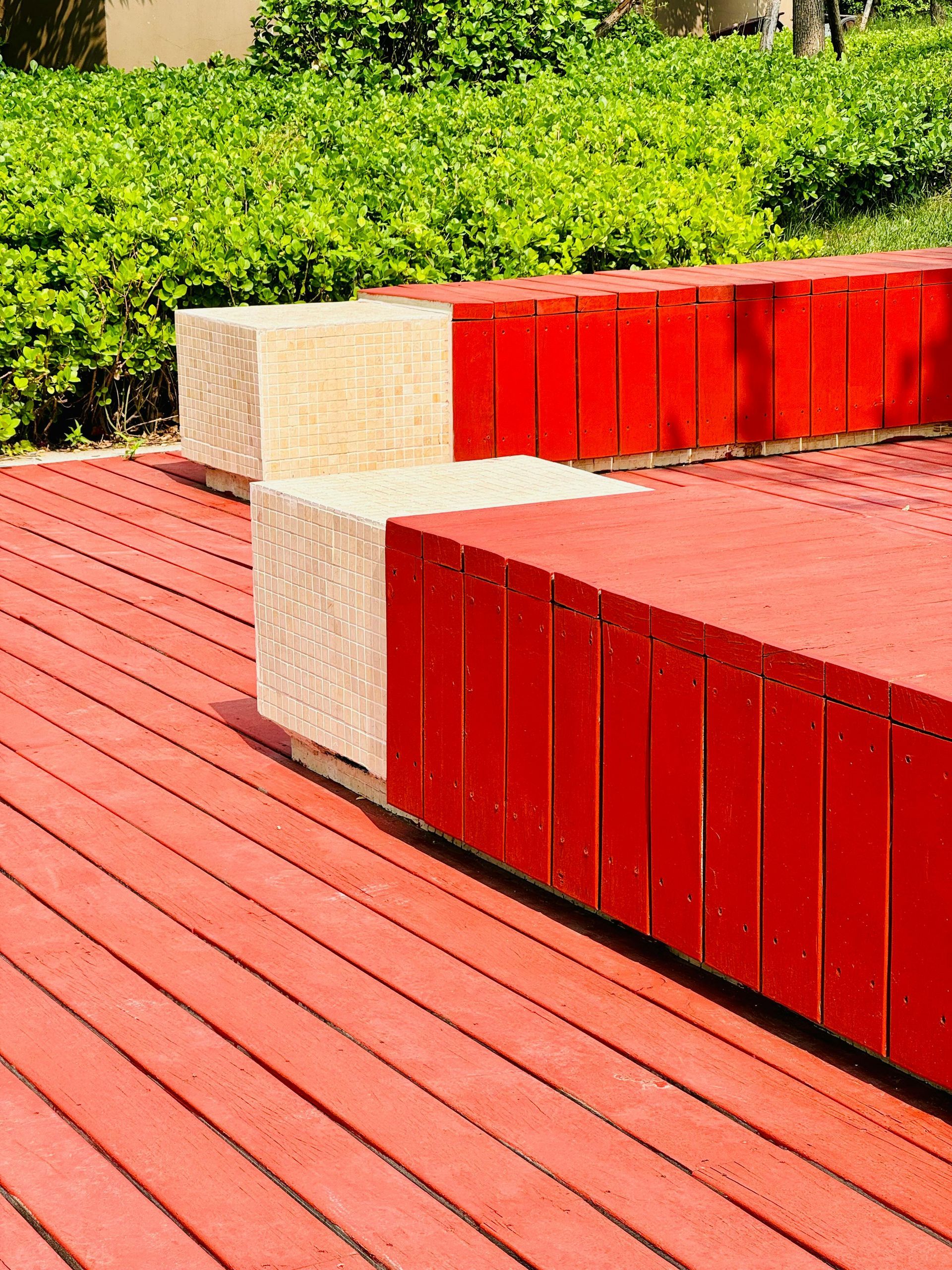 Red benches on a wooden deck beside green shrubs in bright sunlight