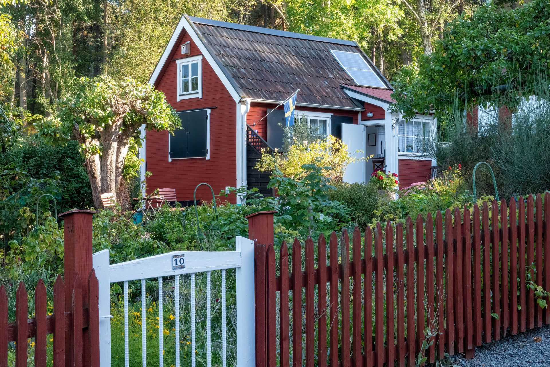 Red house with white trim behind a red fence and white gate, surrounded by trees.