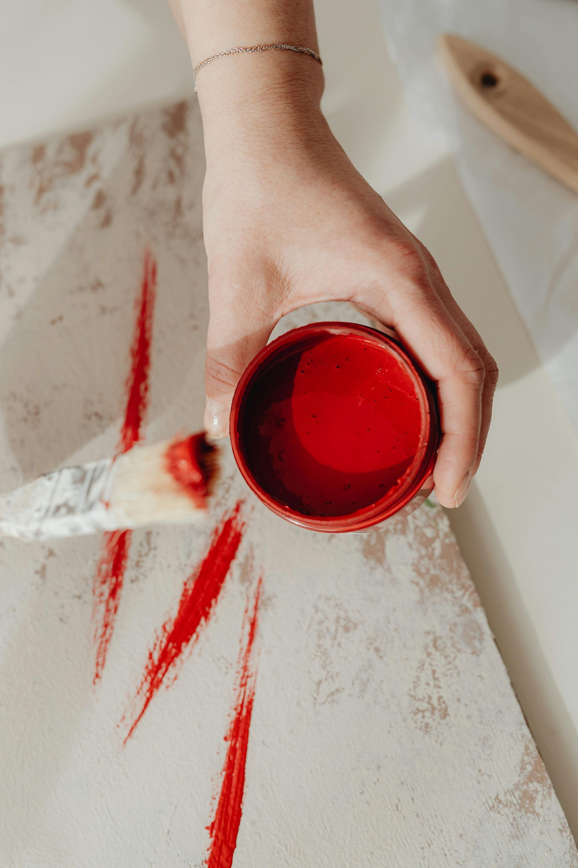 Hand pouring red paint from a cup onto a white wooden surface, creating streaks