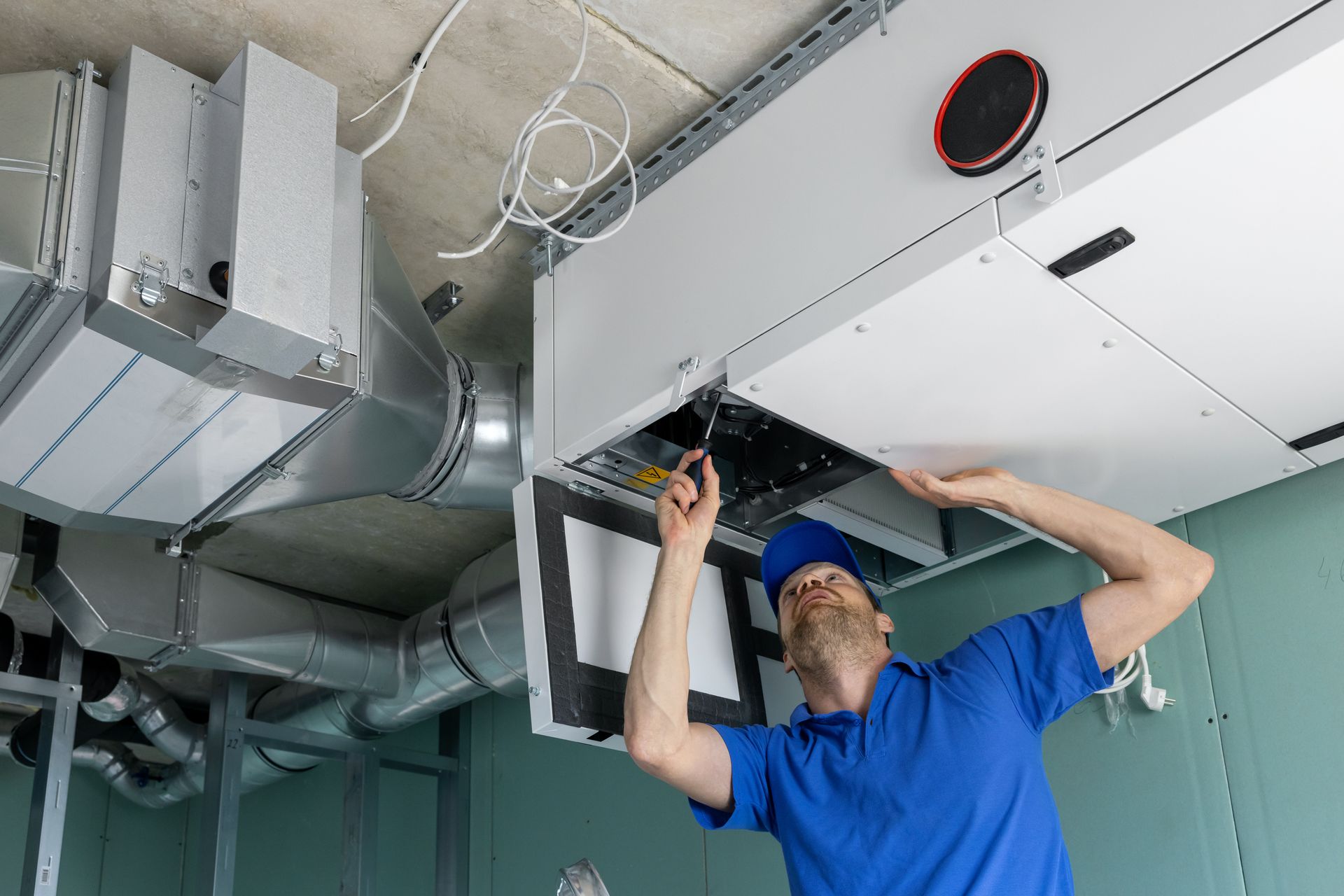 A technician in a blue shirt works on an open ventilation unit mounted on a ceiling.