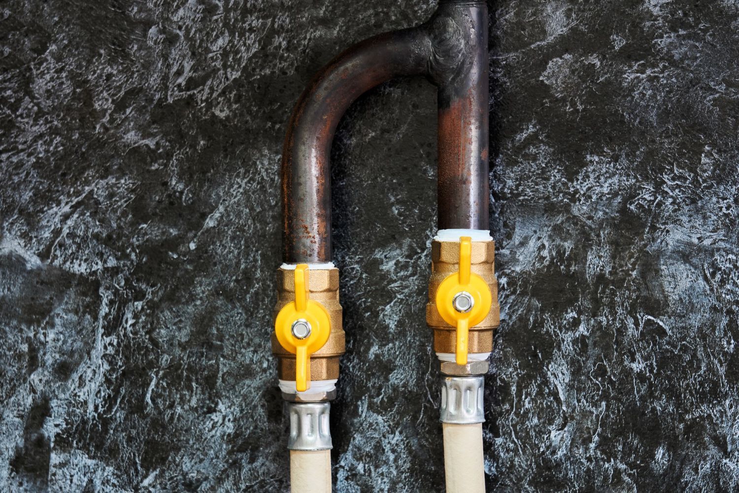 Two copper pipes with bright yellow shut-off valves connected to hoses against a dark, mottled background.