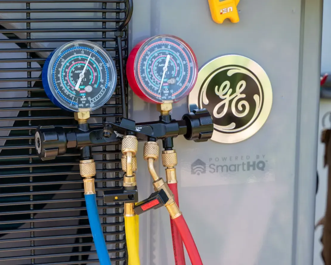 Three HVAC manifold gauges with blue, yellow, and red hoses on a GE unit panel.