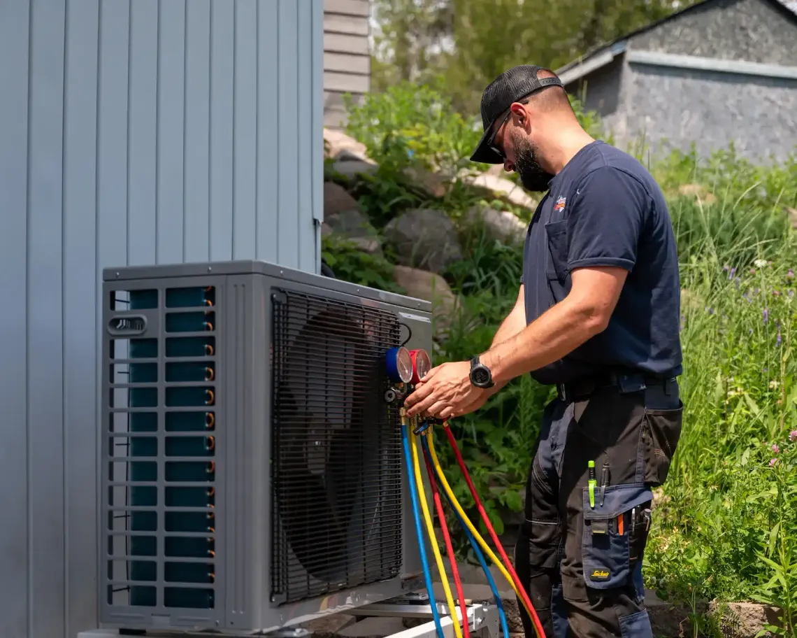 Technician servicing an outdoor HVAC unit, holding colorful refrigerant hoses beside a house