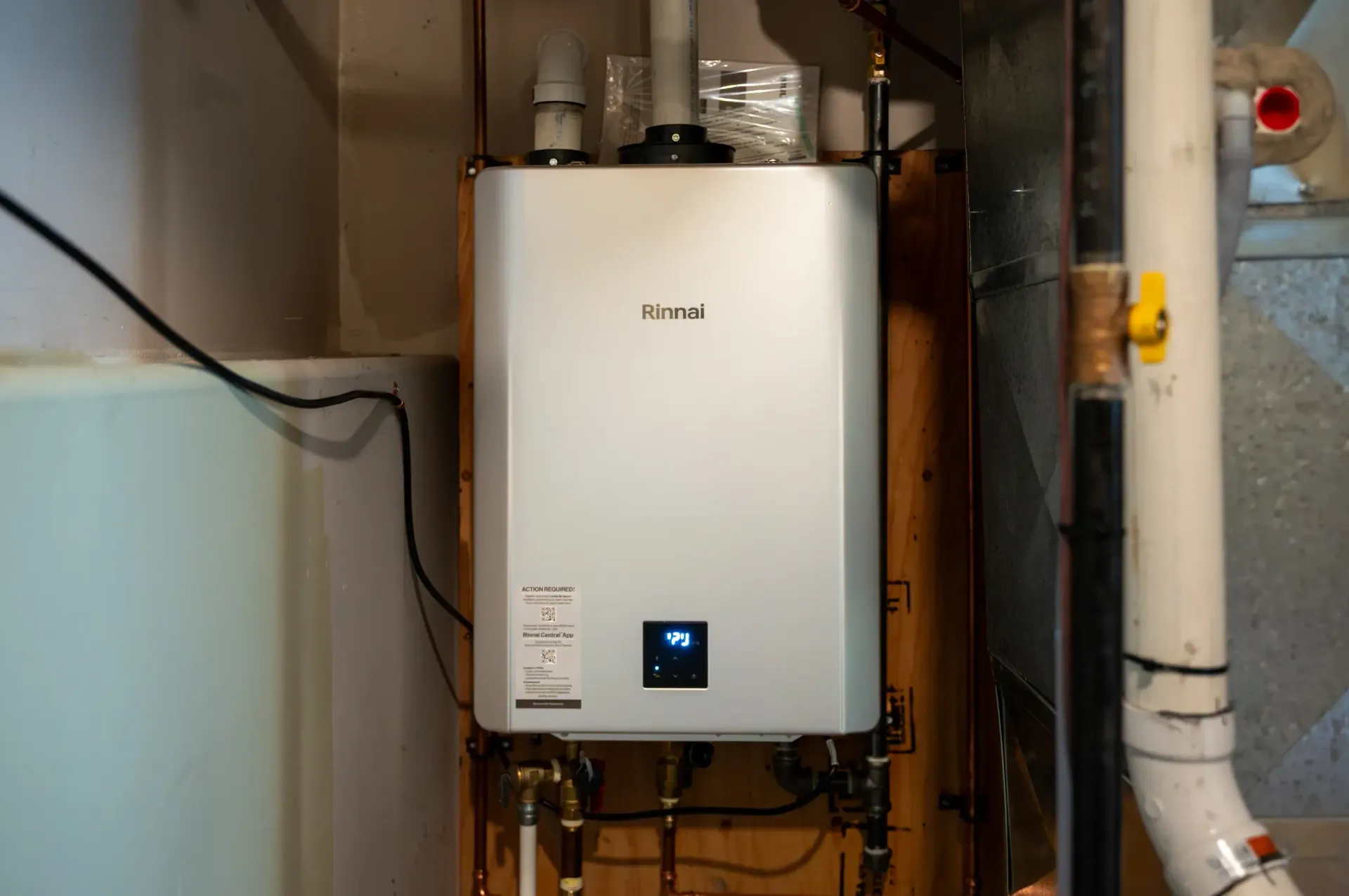 Wall-mounted white water heater with blue display in a utility closet, surrounded by pipes and wiring