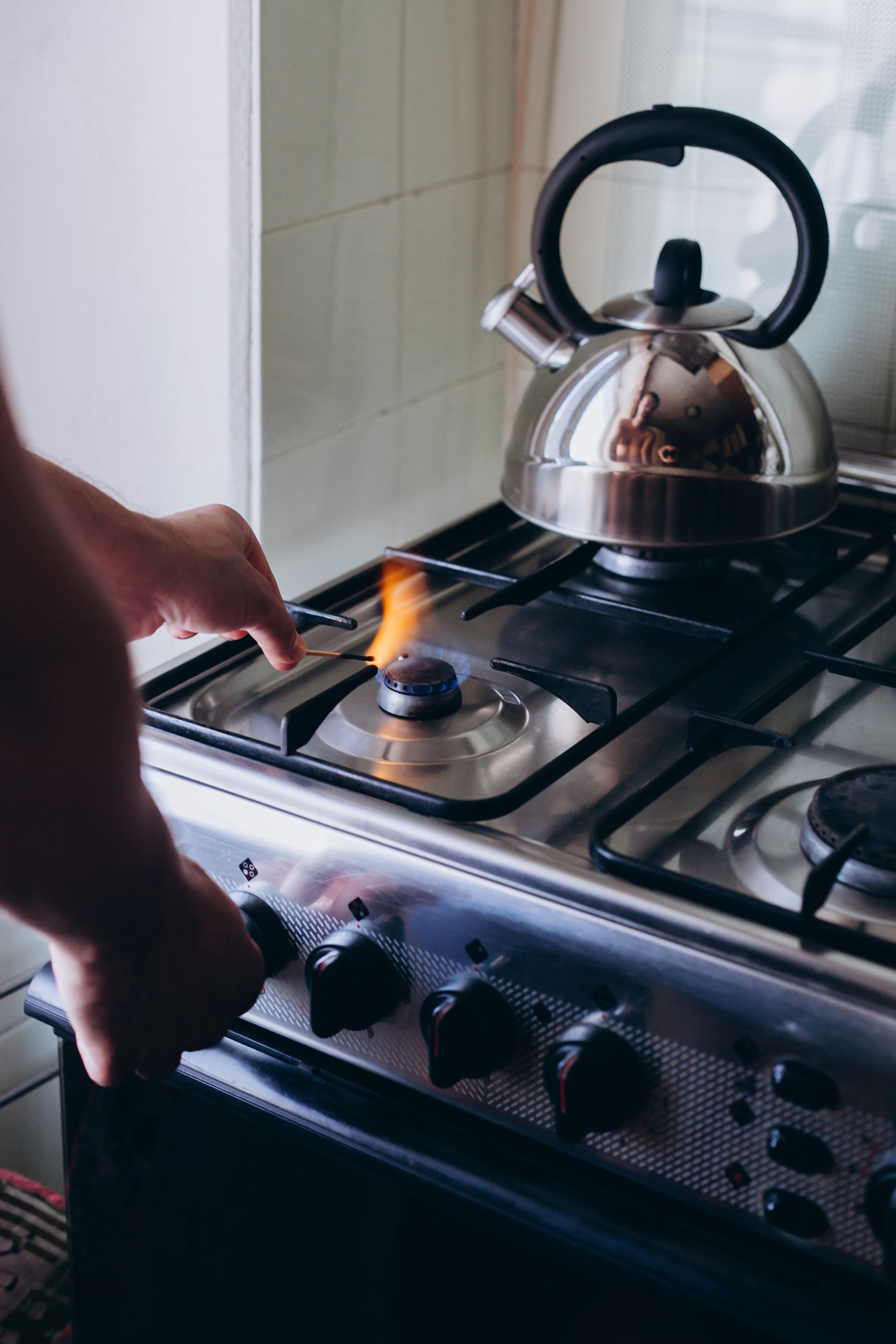 Person lighting a gas stove burner beside a silver kettle in a kitchen