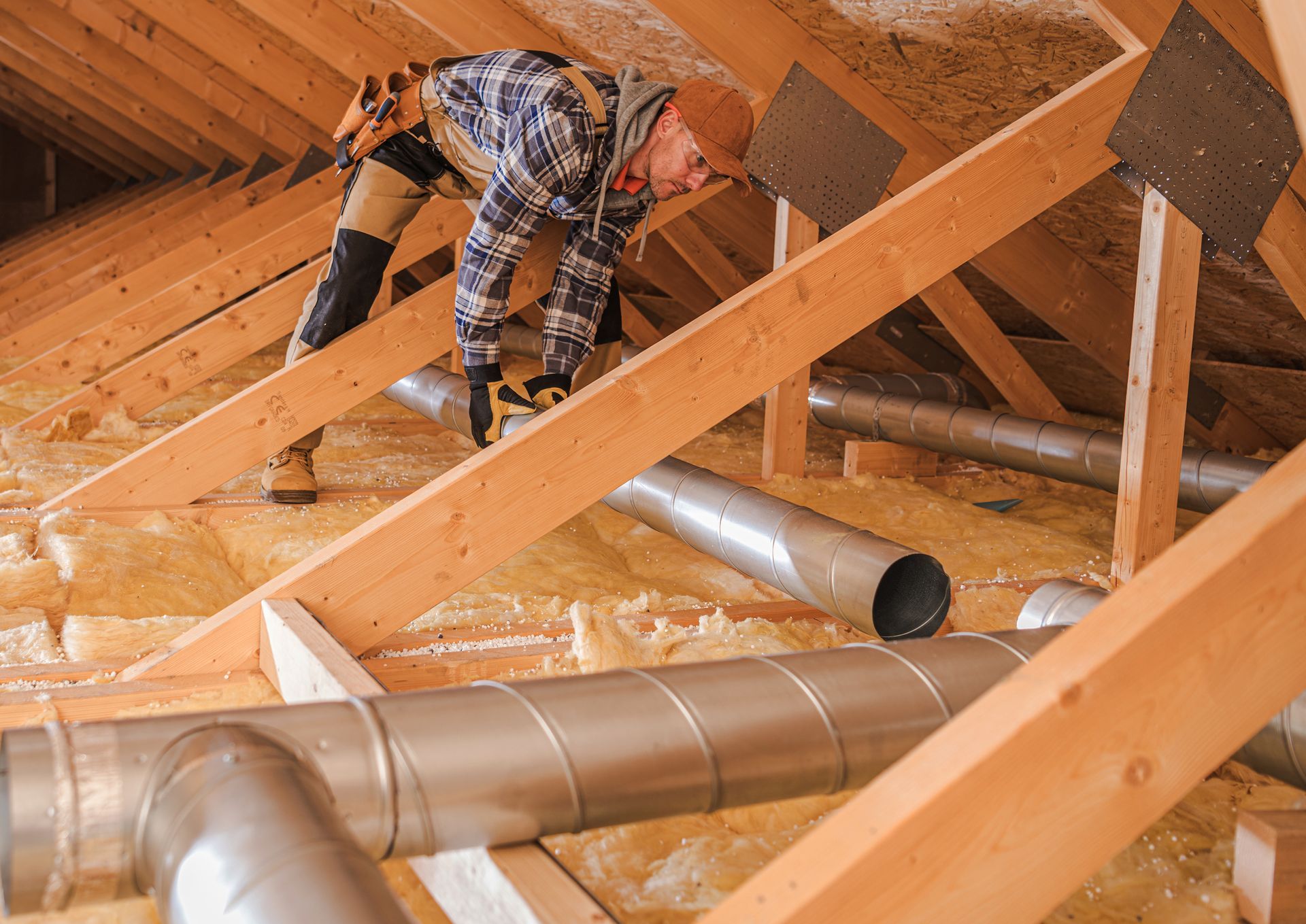 Worker installing ductwork in an attic with exposed wooden beams and insulation.