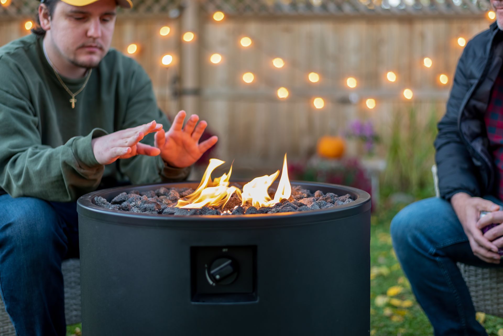 Person warming hands over a fire pit outdoors at dusk, with another person seated nearby