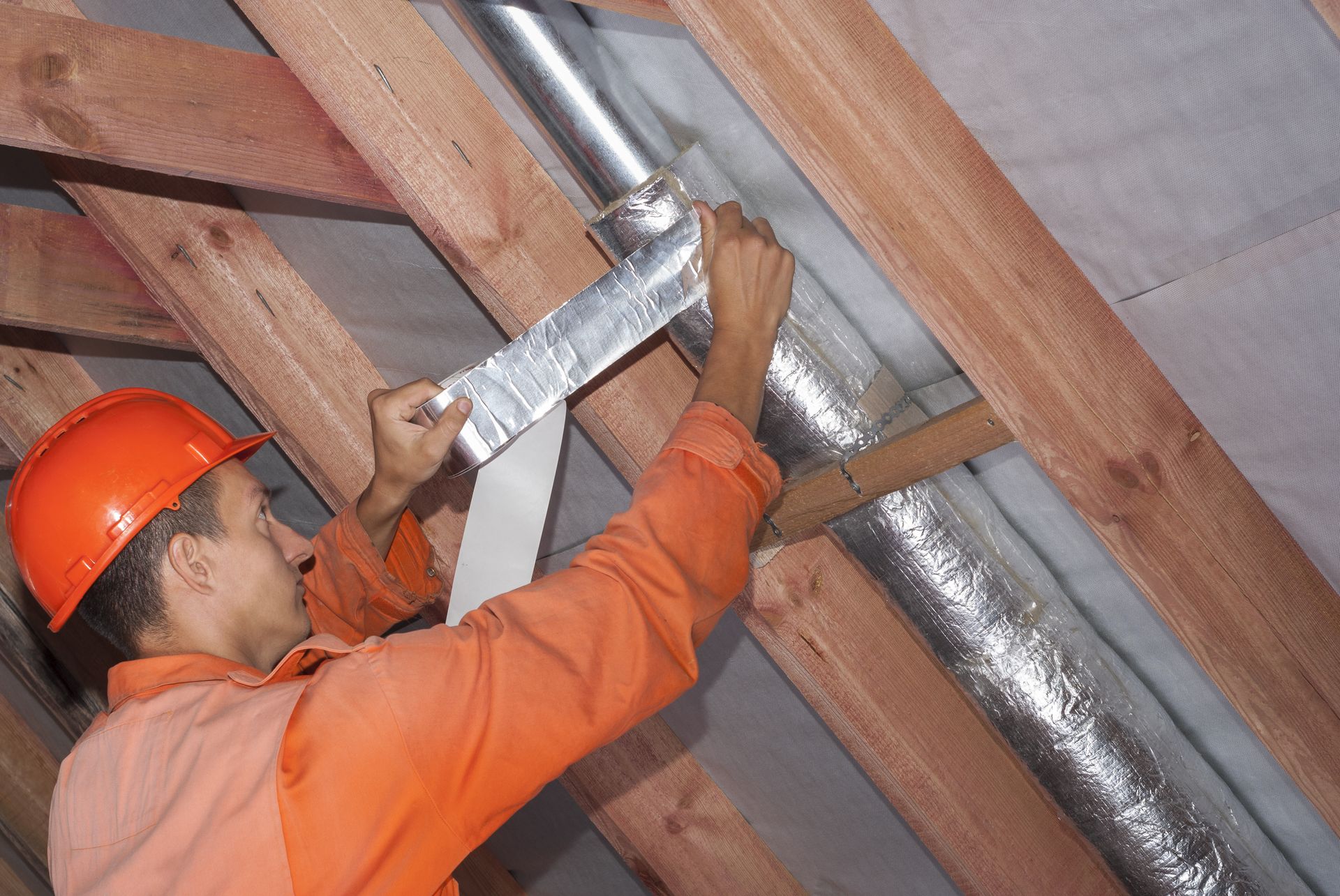 Worker in orange hard hat installing ductwork in an attic space