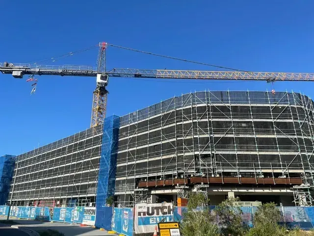 Construction site with crane, building covered in scaffolding and safety netting, blue sky.